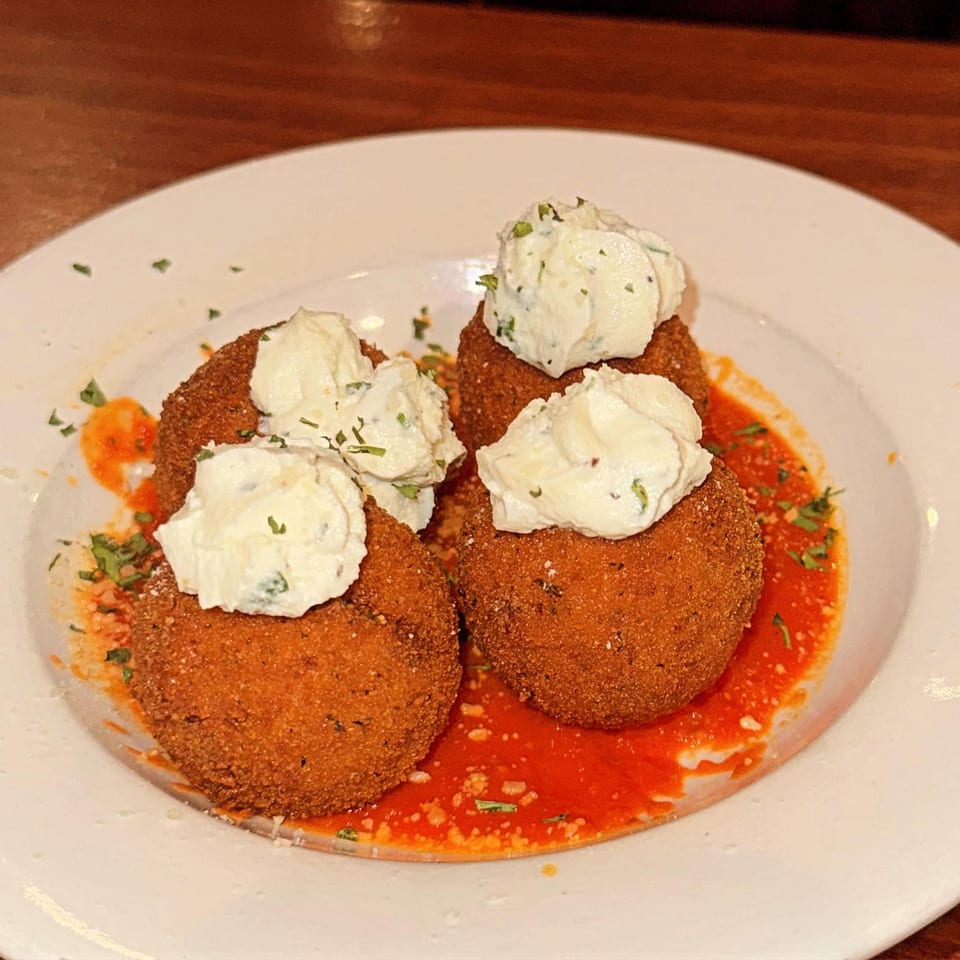 Four golden-brown arancini balls topped with creamy white cheese and parsley, resting in red sauce.