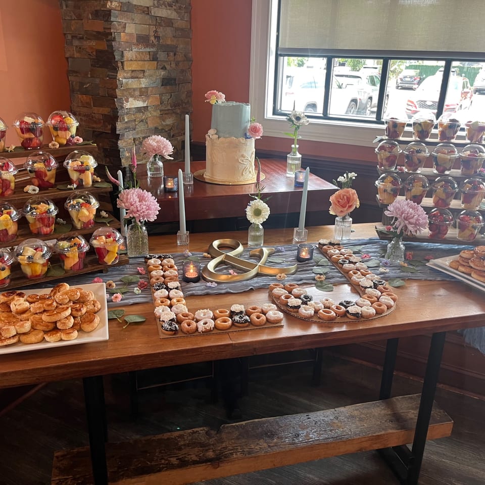 A dessert table with a two-tier cake, fruit cups, mini donuts, and pastries, decorated with flowers.