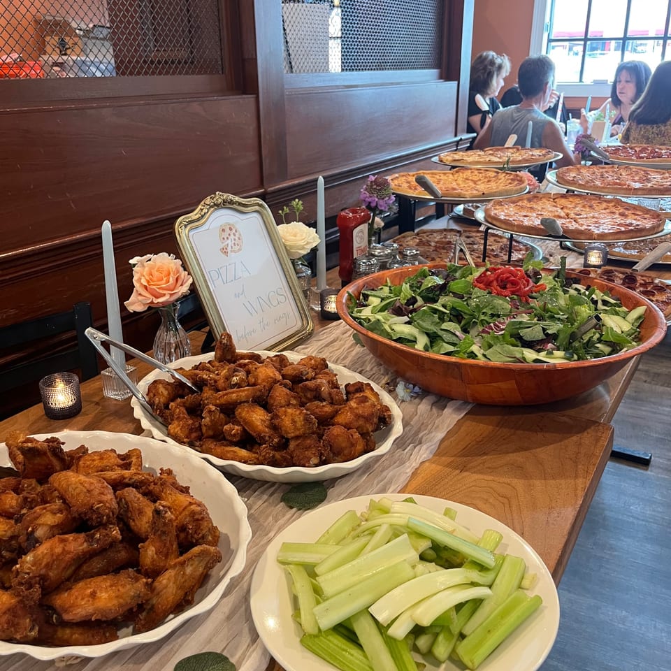 A buffet table with Nanuet Restaurant's pizza, wings, salad, and celery sticks, with diners in the background.